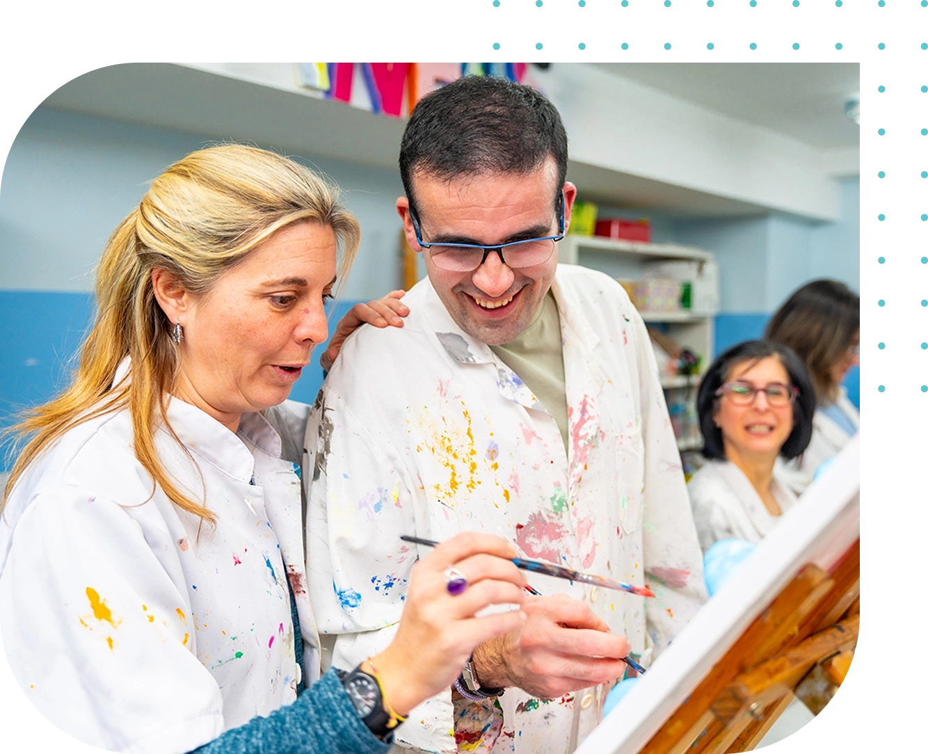 Two people wearing paint-splattered lab coats enjoy painting in a classroom, supported by Disabilities Support Services, while a third person sits and observes in the background. Two people wearing paint-splattered lab coats enjoy painting in a classroom, supported by Disabilities Support Services, while a third person sits and observes in the background.
